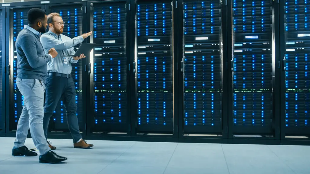 Two engineers discuss data quality management as they walk by a wall full of servers.